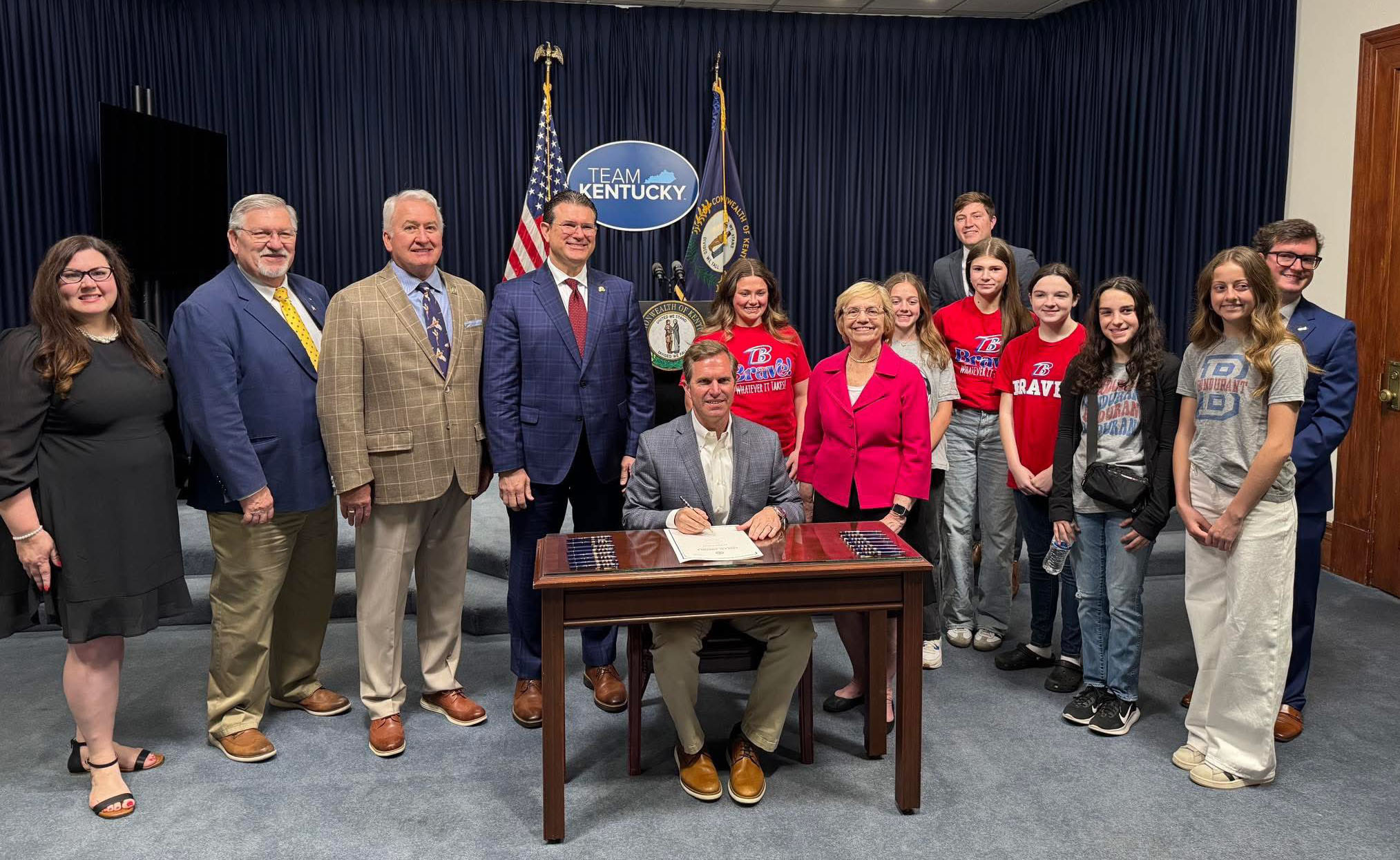 image of a group of adults smiling at the camera in front of a blue curtain. One man, Kentucky Governor Andy Beshear is sitting at a desk with the signed House Bill 190. 